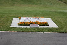 A grave marked by an angled slab of marble engraved with the names of Diefenbaker and his wife and surrounded by plantings of small marigolds, and an additional plaque in bronze. Beyond it a lawn stretches away.